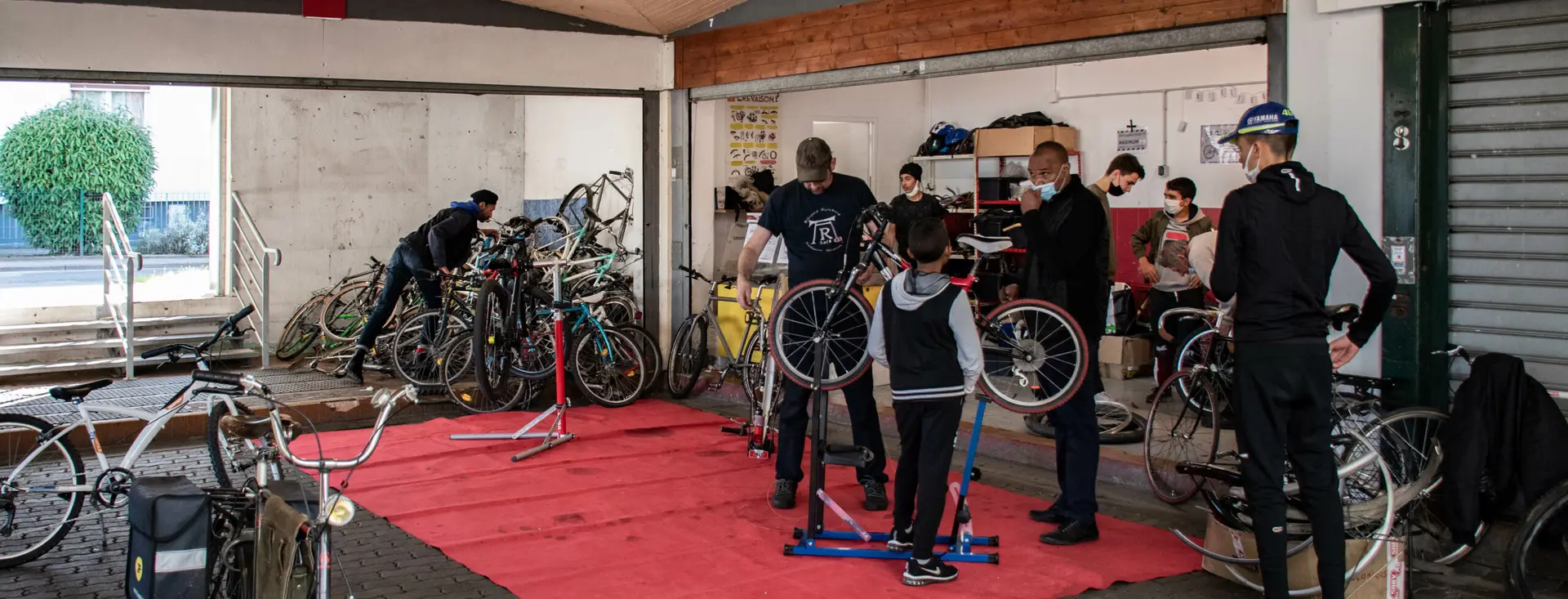 Une salle d’atelier remplie de vélos sur pieds de réparation, une vingtaine de personnes au travail.